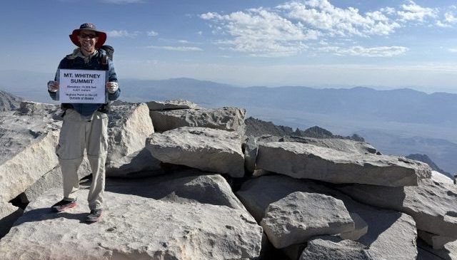 A person standing on the summit of Mt. Whitney, holding a sign marking the elevation and location. The background features rocky terrain and distant mountains under a clear sky.