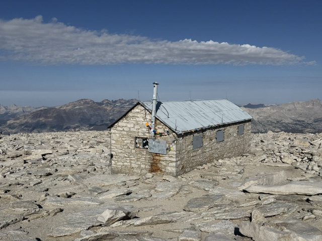 A stone cabin with a metal roof situated on a rocky mountain landscape under a clear blue sky.