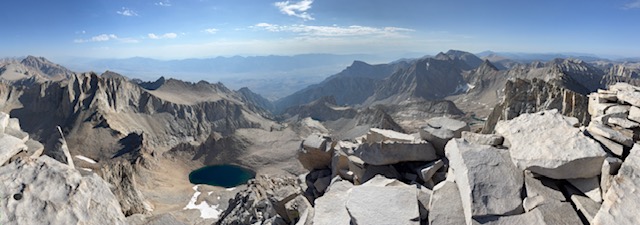 Panoramic view from a mountain summit showcasing rugged terrain, distant peaks, and a blue lake surrounded by rocky slopes under a clear sky.