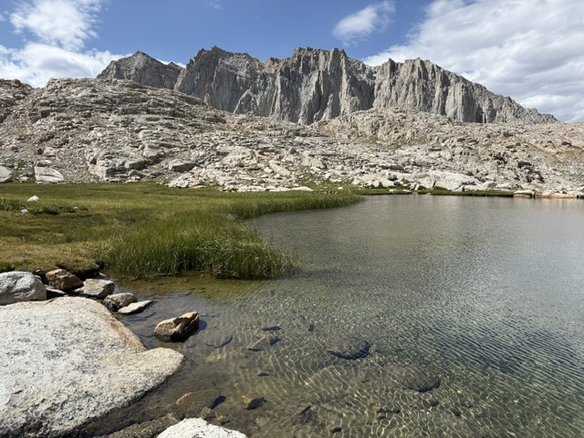 A tranquil mountain lake surrounded by rocky terrain and towering cliffs, with clear water reflecting the sky and patches of green grass along the shore.