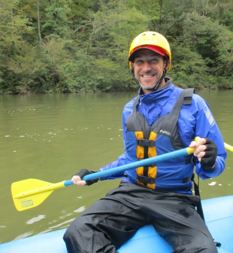 A smiling man wearing a helmet and a blue waterproof jacket is sitting in a raft, holding a yellow paddle, with lush greenery and water in the background.