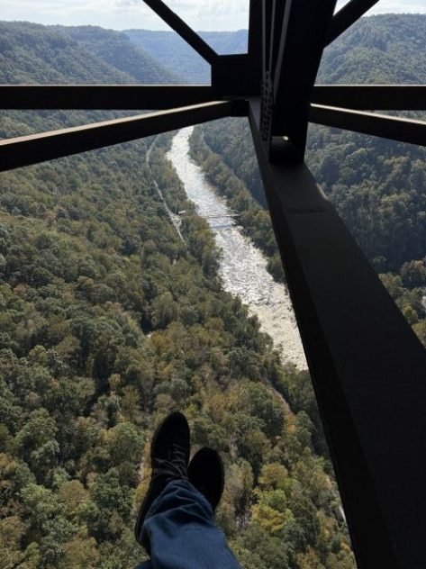 View from above a river valley, showing a winding river and dense tree cover, with a person's feet visible in the foreground.